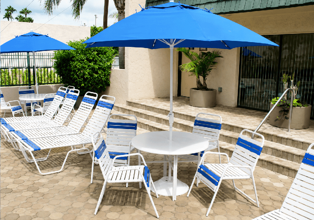 Commercial pool deck featuring white and royal blue vinyl strap dining chairs, round fiberglass tables, and royal blue Frankford market umbrellas creating a shaded outdoor dining area.